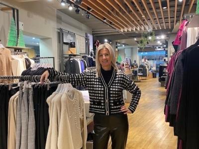 Vrouw in stijlvolle kleding poseert in een goed verlichte kledingwinkel naast een rek met modeartikelen.