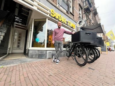 Man leunt tegen bakfiets voor Sushi Time winkel in stadsstraat met terras en fietsparkeren voor de deur.