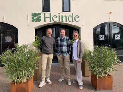 Drie mensen staan voor de ingang van een gebouw met het Heinde-logo, omgeven door groen in plantenbakken.