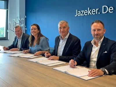 Vier personen ondertekenen documenten aan een tafel met op de achtergrond een blauwe muur en tekst Jazeker. De.