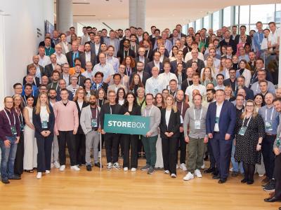 Grote groep mensen poseert in conferentiehal met Storebox-bord, symboliserend samenwerking en groei.