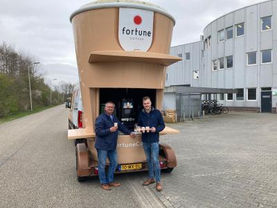 Twee mannen staan bij een groot koffieautomaat op een aanhanger voor een bedrijfsgebouw, met koffie in hun handen.