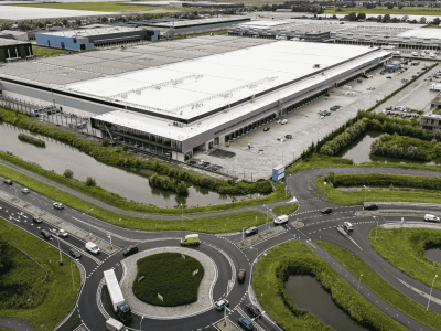Luchtfoto van een groot distributiecentrum omgeven door wegen, parkeerplaatsen en omliggende groene gebieden.