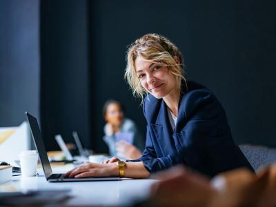 Vrouw in kantooromgeving werkt aan een laptop, glimlachend naar de camera met collega op de achtergrond.