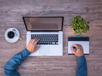 Persoon werkt aan een laptop met een tekentablet, een plantje en een kop koffie op een houten bureau.