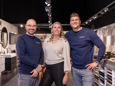 Drie mensen poseren in een showroomomgeving bij Sanidirect, met sanitairproducten op de achtergrond.