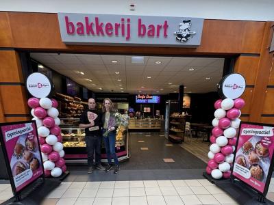 Twee ondernemers staan voor hun Bakker Bart winkel met bloemen en een trofee, omringd door ballonnen en reclameborden.