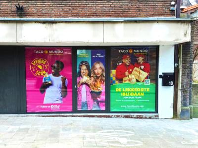 Gevel met kleurrijke posters van Taco Mundo, aankondiging van een nieuwe vestiging in Heerlen met wervingsoproep.