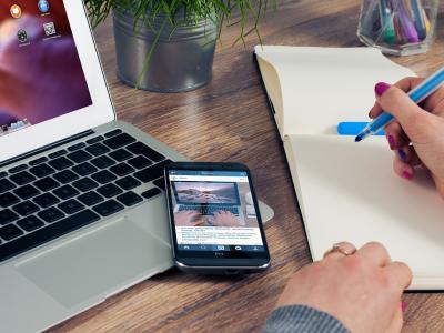 Hand schrijft in notitieboek naast laptop en smartphone op een bureau.