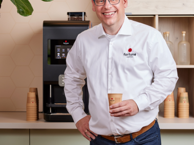 Man in Fortune Coffee-uniform met papieren beker bij koffiezetapparaat, omringd door koffiebekers en planten.