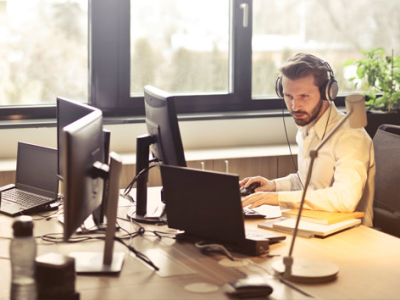 Medewerker met koptelefoon werkt geconcentreerd achter meerdere computerschermen in een licht kantoor.