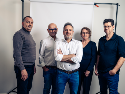 Vijf volwassenen staan professioneel opgesteld voor een witte achtergrond in een fotostudio-omgeving.
