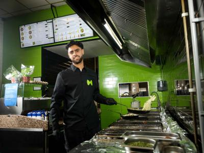Man in een keuken van een restaurantfranchise bakt voedsel terwijl hij naar de camera kijkt, met menu's op de achtergrond.