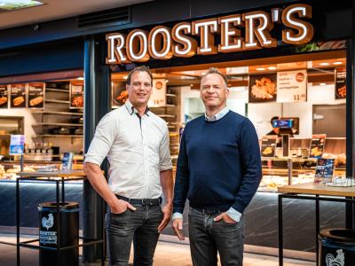 Twee mannen staan voor een Rooster’s vestiging met een uitnodigend interieur en menuborden.