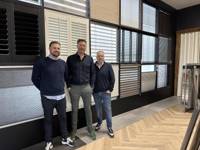 Drie mannen staan voor een wand met diverse gordijn- en jaloezieënstalen in een showroom.