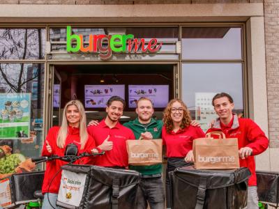 Medewerkers van Burgerme poseren met bezorgfietsen en tassen voor de winkel, klaar voor maaltijdlevering.