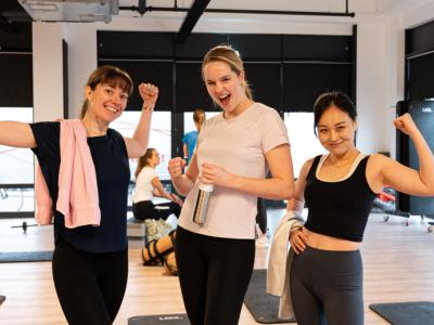 Drie lachende vrouwen tonen spierballen in een fitnessstudio met yoga- en trainingsmatjes op de vloer.