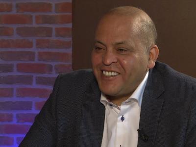 Man in donkerblauw pak lacht voor een muur van rode bakstenen.