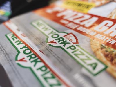 Folder van New York Pizza met promotietekst en deel van een pizza-afbeelding.