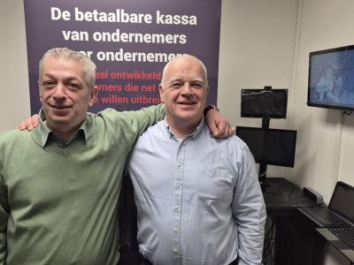 Twee mannen poseren in een kantoor met slimme kassasystemen en promotiemateriaal op de achtergrond.