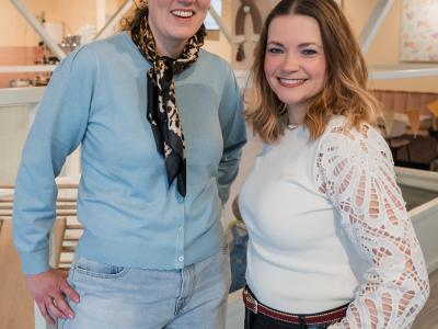 Twee glimlachende vrouwen poseren in een gezellig ingerichte ruimte met decoraties en planten.