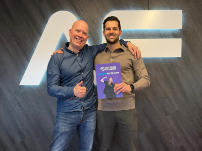 Twee mannen poseren voor het Anytime Fitness-logo, één houdt een welkomstbrochure vast.