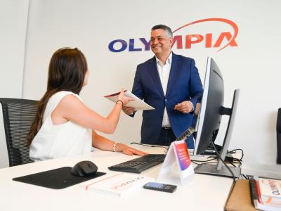 Man in blauw pak overhandigt een map aan zittende vrouw in een kantoor met Olympia-logo op de achtergrond.