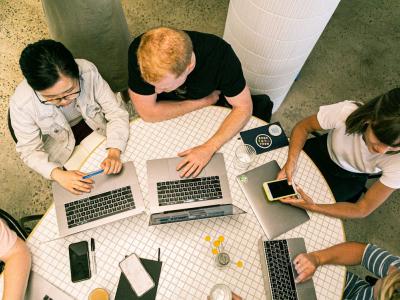 Team van vijf personen werkt samen aan tafel met laptops en notitieboekjes, focus op digitale samenwerking.