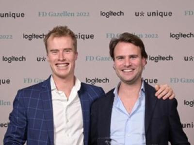 Twee mannen in zakelijke kleding poseren samen op een evenement met merken op de achtergrond.