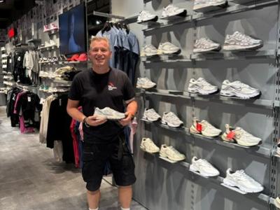 Man in sportkleding houdt een sneaker vast in een moderne winkel met een wand vol damesschoenen en kledingrekken.
