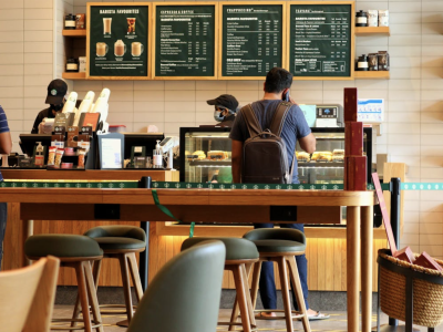Klanten bestellen koffie en snacks bij de balie van een franchise café, met menu's zichtbaar op de achtergrond.