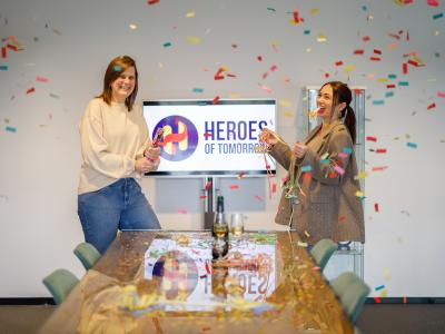Twee mensen vieren feest in een kantoor met confetti, terwijl op de achtergrond een scherm met Heroes of Tomorrow-logo staat.