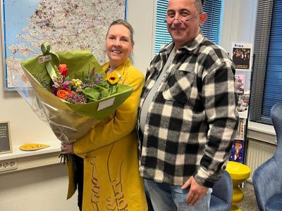 Vrouw in gele jas met bloemen en man in geruite jas poseren voor een kaart van Nederland in een kantooromgeving.