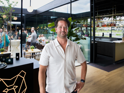 Man in wit overhemd staat in modern restaurantinterieur met glazen wanden en mensen op achtergrond.