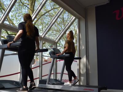 Twee personen sporten op loopbanden bij grote ramen in een sportschoolomgeving.