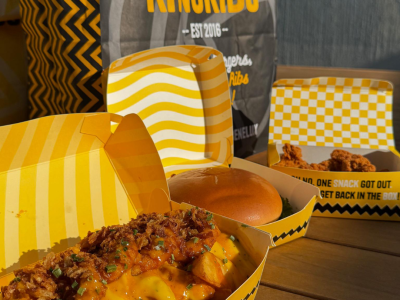 Kingribs take-out met een hamburger, friet met saus en gefrituurde kip in gele verpakkingen op een houten tafel.