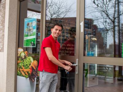 Man in rood shirt opent de deur van een Burgerme-vestiging met het logo zichtbaar boven de ingang.