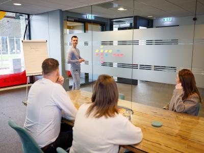 Mensen in een vergaderruimte overleggen terwijl een spreker hun aandacht trekt met een flip-over en post-its op glazen wand.