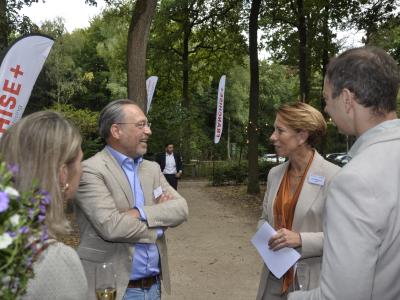 Deelnemers netwerken op de Franchise+ BBQ in een bosrijke omgeving met vlaggen en hapjes in de hand.