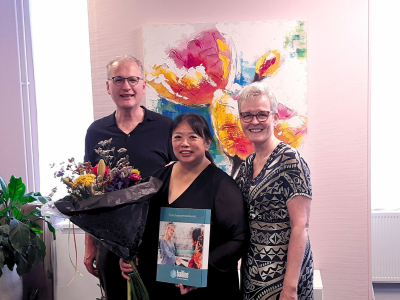Drie mensen poseren binnen met een bloemenboeket en een Bailine-franchiseovereenkomst, met kleurrijke kunst op de achtergr...