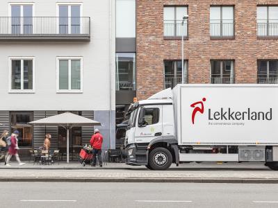 Lekkerland vrachtwagen levert producten aan een locatie met terrassen waar mensen lopen en een medewerker werkt.