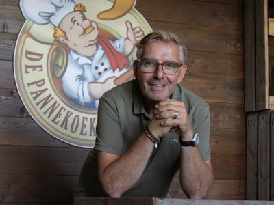 Man leunt glimlachend op houten balie voor een logo van De Pannekoekenbakker op houten achtergrond.