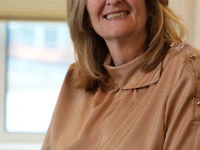 Vrouw met blond haar en een lichtbruine blouse glimlacht naar de camera, zittend voor een raam met gesloten lamellen.