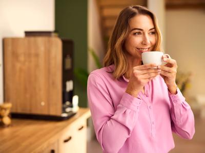 Vrouw glimlacht terwijl ze een kopje koffie drinkt in een sfeervolle kantooromgeving met een koffiemachine op de achtergrond.