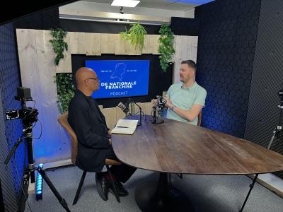 Twee mensen in gesprek aan een tafel in een opnamestudio voor De Nationale Franchise Podcast.