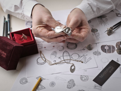 Een persoon inspecteert sieraden met een loep boven schetsen van sieradenontwerpen op een tafel.