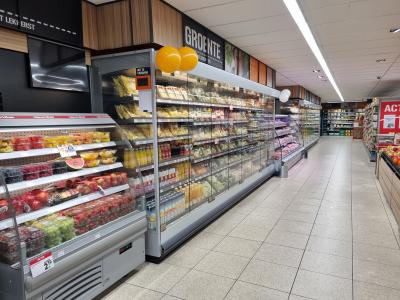 Supermarktgang met koelvitrines vol gesneden fruit, groenten, sapjes en andere gekoelde producten, versierd met ballonnen.