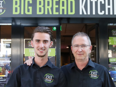 Twee medewerkers in BB Kitchen-uniform poseren voor de ingang van een Big Bread Kitchen-filiaal.