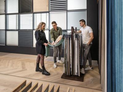 Twee mannen en een vrouw bekijken gordijnstalen in een showroom met verschillende stoffen aan de wand.