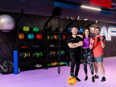 Drie mensen poseren in een fitnessruimte met sportuitrusting op de achtergrond.